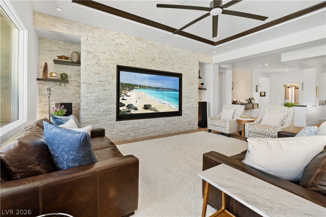 Great room at 3084 Red Arrow Dr with a full-height white stacked-stone media wall, recessed TV, coffered ceiling with dark wood beams, two leather sofas, and printed accent chairs