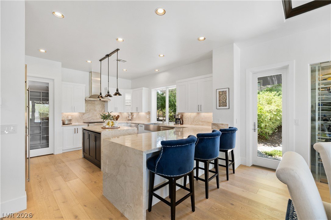 Chef's kitchen at 3084 Red Arrow Dr with an oversized marble waterfall island, three deep-blue velvet counter stools, white cabinetry, brushed brass pendants, and a door to the pool courtyard